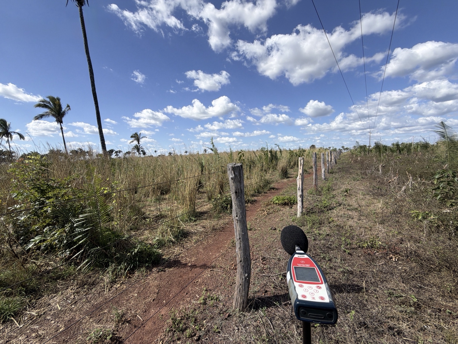 Medidor de nível sonoro instalado em campo próximo a cerca e vegetação.